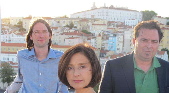 Drei Personen stehen vor einer mediterranen Stadtlandschaft in warmem Licht. Eine Frau in buntem Kleid blickt ernst in die Kamera, zwei Männer stehen dahinter und schauen ebenfalls nach vorn.
