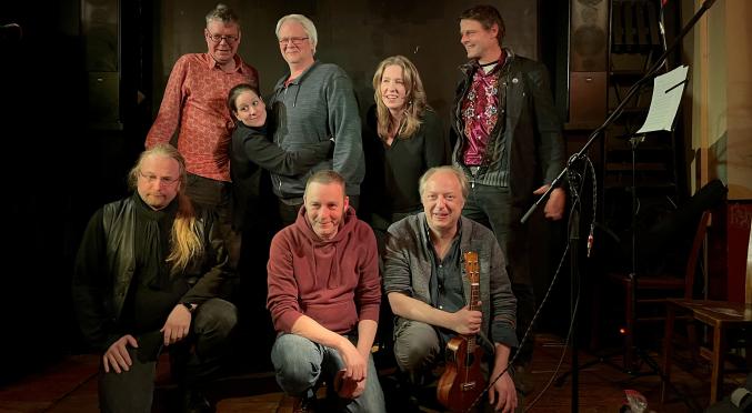 Acht Menschen posieren fröhlich auf einer kleinen Bühne für ein Gruppenfoto. Drei Personen knien vorn, einer hält eine Ukulele, fünf stehen dahinter. Die Stimmung ist warm und freundschaftlich.