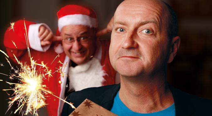 Ein Mann mit ernstem Blick hält ein Geschenk, im Hintergrund hält ein fröhlicher Mann im Weihnachtsmannkostüm die Ohren zu, während eine Wunderkerze hell aufleuchtet.