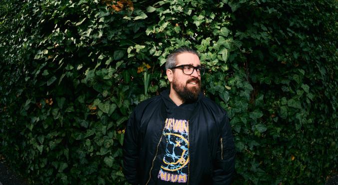 Ein Mann mit Bart und Brille steht vor einer dichten grünen Efeuhecke. Er trägt eine schwarze Jacke und ein Nirvana-Shirt und blickt lächelnd zur Seite.