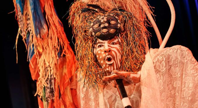Person mit bemaltem Gesicht, bunter Masken-Kopfbedeckung und Spitzengewand singt oder spricht expressiv ins Mikrofon bei einer Bühnenperformance mit farbenfrohem Dekor.