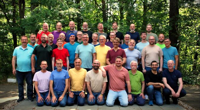 Eine große Gruppe von Männern in bunten T-Shirts steht und kniet lachend im Grünen für ein Gruppenfoto im Wald.