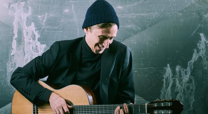 Ein junger Mann mit dunklem Anzug und Mütze sitzt lächelnd auf einer Bank und spielt Gitarre vor einer rauen, industriellen Wand.