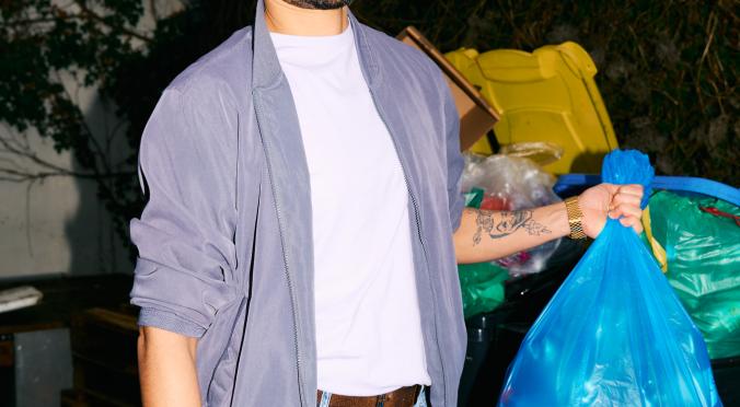 Ein Mann mit ernster Miene steht vor Mülltonnen und hält einen blauen Müllsack. Er trägt Jeans, weißes T-Shirt und eine graue Jacke. Im Hintergrund ist dichte Vegetation.