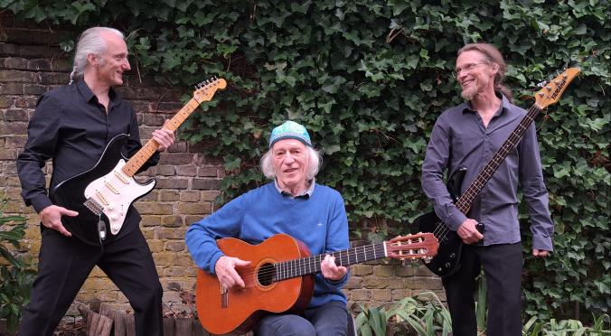 Drei ältere Männer stehen vor einer efeubewachsenen Mauer. Zwei stehen mit E-Gitarren, einer sitzt mit Akustikgitarre. Alle lächeln, die Stimmung ist entspannt und musikalisch.