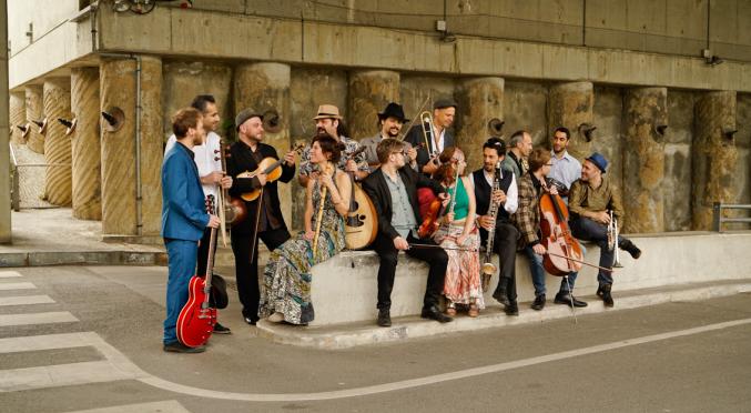 Eine Gruppe von Musikern sitzt und steht auf einem Bordstein vor einer Betonwand, spielt verschiedene Instrumente und lacht zusammen. Es wirkt wie ein fröhliches Straßenkonzert.