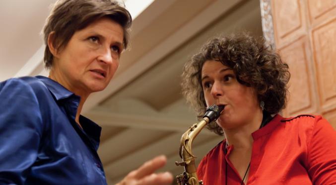 Zwei Personen stehen zusammen: Eine in einem blauen Hemd spricht, während die andere in einem roten Hemd konzentriert Saxophon spielt. Die Szene wirkt wie eine Probe oder Unterricht.