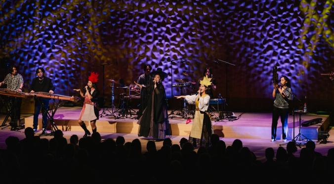 Achtköpfige Band auf Bühne mit traditionell-kreativen Outfits, bunter Beleuchtung und koreanischen Instrumenten, singend und musizierend vor Publikum.