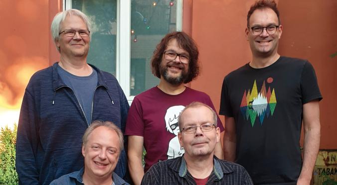 Fünf Männer stehen und sitzen lächelnd in einem Garten vor einer orangefarbenen Wand mit Fenster. Es ist ein Gruppenfoto in entspannter, freundlicher Atmosphäre.