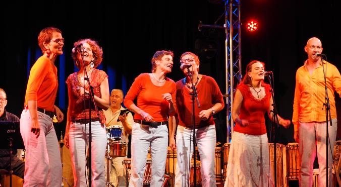 echs Personen in roten Oberteilen und weißen Hosen singen fröhlich auf einer Bühne. Im Hintergrund spielen Musiker Percussion-Instrumente. Bunte Lichter beleuchten die Szene.