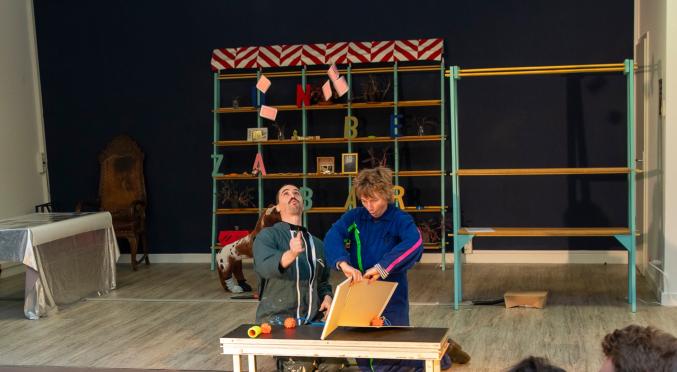 In einem Raum führen zwei Personen eine Theateraufführung auf. Sie sitzen auf einer Bank: eine in dunkler Kleidung und die andere im blauen Overall. Im Hintergrund sind Regale und eine mit Flaggen geschmückte, dunkle Wand. Zuschauer sind im Vordergrund sichtbar.