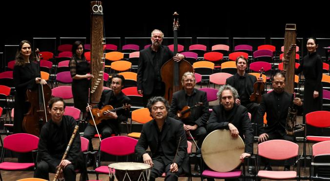 Zehn Musiker in schwarzer Kleidung posieren mit traditionellen und modernen Instrumenten. Sie sitzen und stehen zwischen bunten Stühlen auf einer Bühne. Die Atmosphäre wirkt professionell und kulturell.