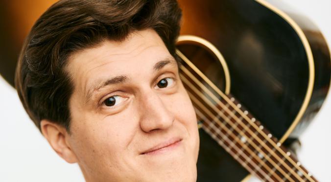 Ein junger Mann mit braunen, gepflegten Haaren und einem fröhlichen Lächeln trägt ein helles T-Shirt und hält eine Gitarre auf der Schulter. Er wirkt entspannt und freundlich.