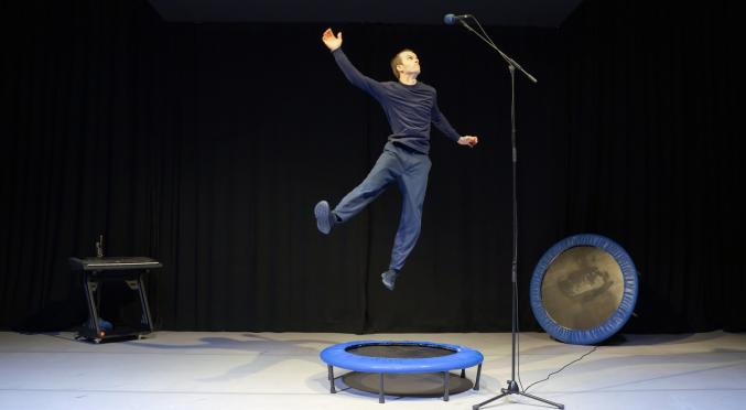 Ein Mann springt auf einem kleinen blauen Trampolin in die Luft, hebt einen Arm und steht vor einem Mikrofon auf der Bühne. Im Hintergrund sind ein weiteres Trampolin und ein Keyboard zu sehen.