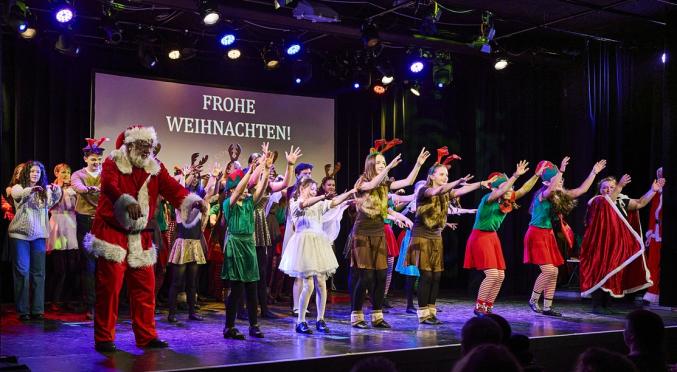 Eine große Gruppe verkleideter Menschen, darunter ein Weihnachtsmann, Elfen und Rentiere, steht auf einer festlich beleuchteten Bühne. Im Hintergrund leuchtet die Botschaft „Frohe Weihnachten!“.