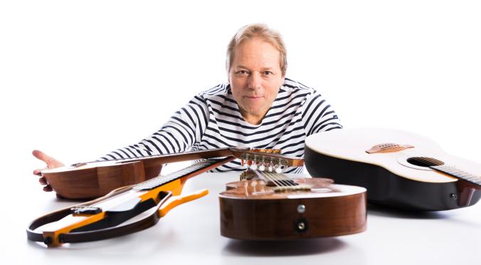 Ein Mann im gestreiften Shirt lehnt nach vorn und schaut in die Kamera. Vor ihm liegen mehrere Gitarren auf einem weißen Tisch, die er mit ausgestreckten Armen umfasst.