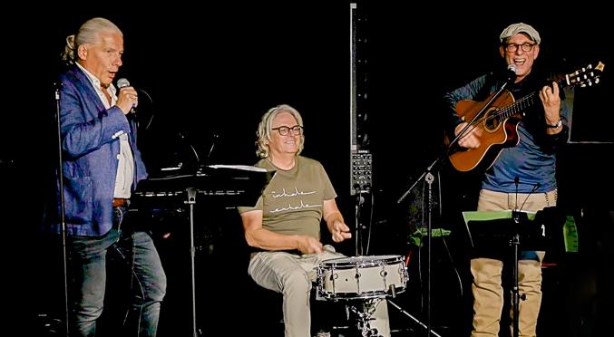 Drei Musiker auf der Bühne. Links mit Notenständer und Gesangsmikrofon, in der Mitte mit einer Snare-Drum und auf der rechten Seite mit einer Gitarre.