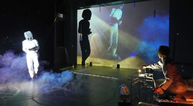Eine Person im weißen Anzug mit Helm steht in Nebel, während rechts jemand an einem Projektortisch arbeitet. Im Hintergrund sind Projektionen von Figuren auf einer großen Leinwand zu sehen.