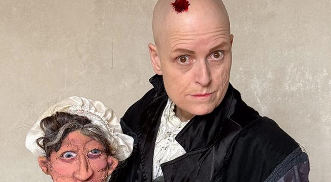 Person mit kahlem Kopf in historischer Robe posiert neben einer Puppenfigur, die wie eine ältere Frau mit Haube und Schürze aussieht.