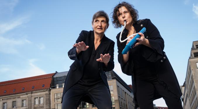 Zwei Frauen in schwarzer Kleidung stehen draußen vor Gebäuden. Eine spielt eine blaue Melodica, die andere gestikuliert mit den Händen, beide blicken konzentriert nach vorn.