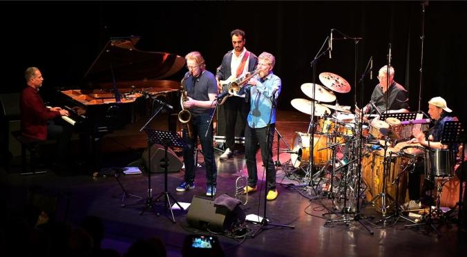 Fünf Musiker auf einer Bühne: Ein Pianist, ein Saxophonist, ein Trompeter, ein Schlagzeuger und ein Percussionist spielen vor Publikum.