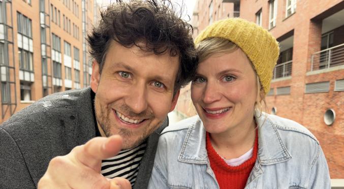 Zwei lächelnde Personen – eine mit gelber Strickmütze und Denim-Jacke, die andere mit dunklem Jackett über gestreiftem Shirt – zeigen auf etwas, im Hintergrund moderne Gebäude.