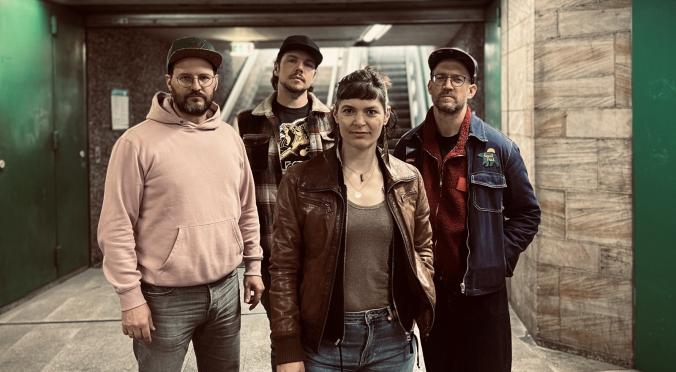 Vier Personen der Band ‚Martha & The Music‘ posieren in einem Tunnel unter einem grünen Logo mit dem Bandnamen. Sie tragen lässige Kleidung wie Lederjacken, Kapuzenpulli und Jeans.