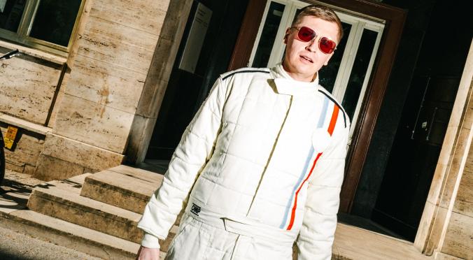 Person in einem weißen Rennfahreranzug mit roten Sonnenbrillen und Helm in der Hand steht auf einer Straße vor einem Gebäude mit Treppen.