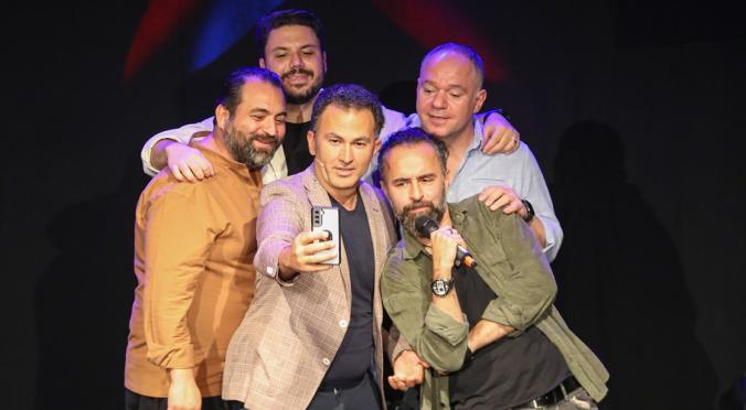 Fünf Männer posieren fröhlich auf einer Bühne: Einer macht ein Selfie, ein anderer hält ein Mikrofon, alle umarmen sich in blauer und roter Bühnenbeleuchtung.