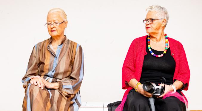 Zwei ältere Frauen sitzen nebeneinander: Eine trägt ein gestreiftes Kleid und Brille, die andere hält eine Kamera, trägt schwarze Kleidung, bunte Kette und robuste Stiefel.