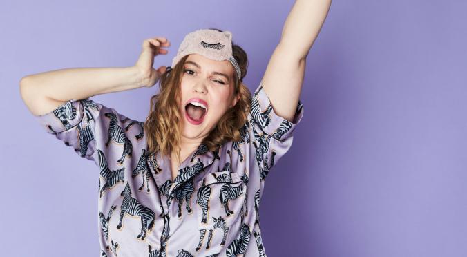Jubelnde Person mit Schlafmaske auf dem Kopf, streckt die Arme nach oben, trägt einen lila Pyjama mit schwarzem Zebramuster und wirkt voller Freude.