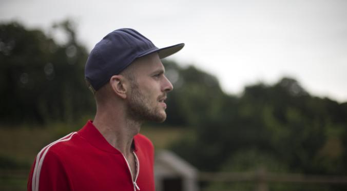 Mann mit blauer Basecap und rotem Sportshirt mit weißen Streifen steht im Profil, blickt nachdenklich in die Ferne, im Hintergrund verschwommene Natur mit Bäumen.