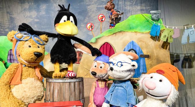 Fünf Stofftiere auf einer Bühne: Ein Bär mit Schwimmbrille, ein Rabe auf einem Fass, ein Kaninchen mit Latzhose, ein Schaf mit Brille und ein Lamm mit Mütze, vor einem Haus und Feuerwehrauto.