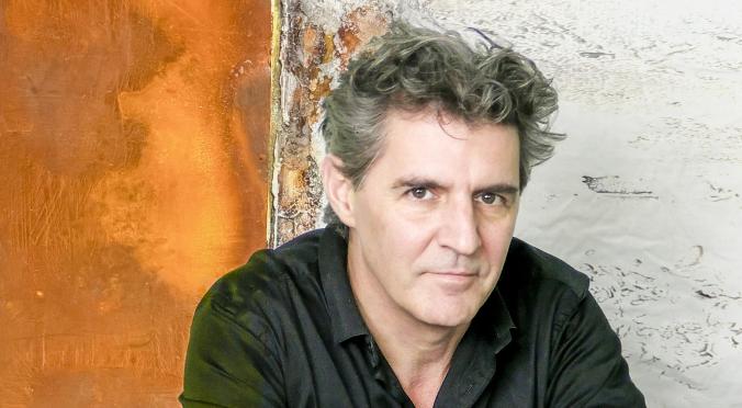 Mann mit graumelierten Haaren und schwarzem Hemd sitzt nachdenklich auf einem glatten Tisch vor einer rostigen Metallwand mit abblätternder Farbe.