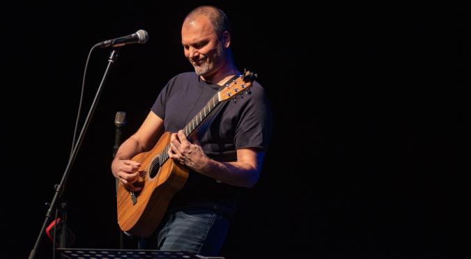 Mann mit Gitarre und dunklem T-Shirt spielt konzentriert auf einer Bühne, ein Mikrofonstativ vor ihm, im Hintergrund dunkel – entspannte, musikalische Atmosphäre.