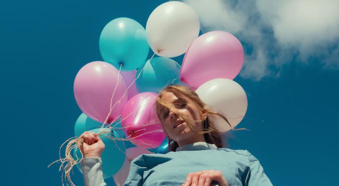 Person lehnt an einer Leiter, hält bunte Luftballons in Rosa, Türkis und Weiß, trägt ein hellblaues Oberteil und wirkt entspannt bei blauem Himmel mit Wolken.