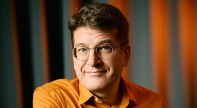 Lächelnder Mann mit Brille und orangefarbenem Hemd lehnt entspannt mit verschränkten Armen auf einem blauen Stuhl vor einem dunklen, vertikal gestreiften Hintergrund.