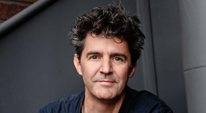 Mann mit struppigen Haaren, dunklem Hemd und Jeans sitzt entspannt vor einer grauen Wand mit Treppengeländer, die Arme lässig auf den Knien abgelegt.