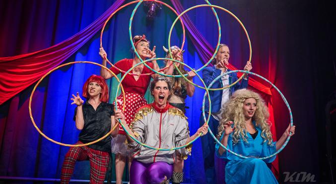 Sechs bunt gekleidete Schauspieler posieren mit Reifen auf einer Bühne: Kostüme in Rot, Blau, Lila, Grau und Perücken, vor blauem Vorhang mit roter Drapierung.