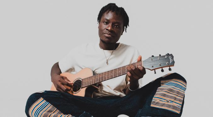 Junger Mann sitzt im Schneidersitz, spielt Gitarre, trägt ein weißes T-Shirt, bunte Hosen mit Streifen und weiße Sneaker mit orange-blauen Akzenten, vor neutralem Hintergrund.