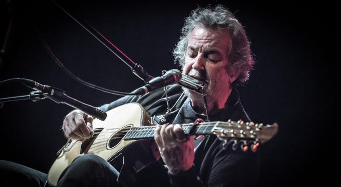 Älterer Musiker mit grauen Haaren spielt konzentriert Gitarre und singt in ein Mikrofon auf einer dunklen Bühne, beleuchtet von rotem Licht.