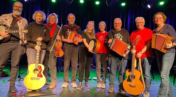 Acht lächelnde Musiker auf einer Bühne: Mit Gitarren, Geige, Akkordeons, Banjo und Trommel, bunt gekleidet, vor blau beleuchtetem Bühnenhintergrund.