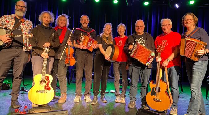 Acht lächelnde Musiker auf einer Bühne: Mit Gitarren, Geige, Akkordeons, Banjo und Trommel, bunt gekleidet, vor blau beleuchtetem Bühnenhintergrund.