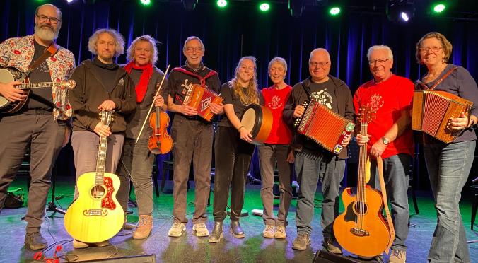 Acht lächelnde Musiker auf einer Bühne: Mit Gitarren, Geige, Akkordeons, Banjo und Trommel, bunt gekleidet, vor blau beleuchtetem Bühnenhintergrund.