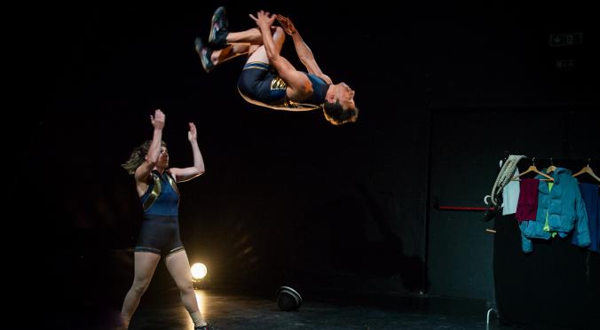 Zwei Akrobaten auf dunkler Bühne: Eine Frau steht mit erhobenen Armen, ein Mann fliegt kopfüber durch die Luft, im Hintergrund eine Person mit Kleiderständer und Seil.