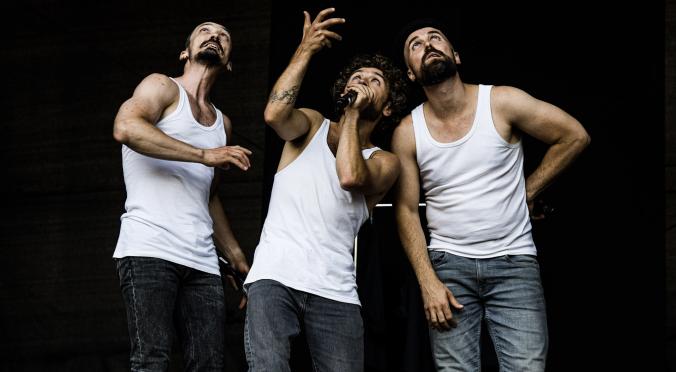 Drei Männer in weißen Unterhemden und Jeans: Der linke streckt Arm aus, der mittlere singt in ein Mikrofon, der rechte lehnt sich lässig zurück, alle vor schwarzem Hintergrund.