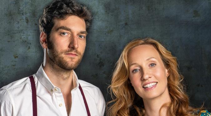 Zwei lächelnde Personen vor metallisch-blauem Hintergrund: Mann mit weißem Hemd und Hosenträgern, Frau mit langem rotblondem Haar in blumigem Oberteil.