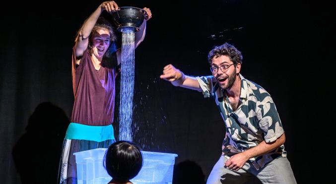 Bühnenbild: Frau gießt Wasser aus einem Sieb in eine durchsichtige Kiste, vor der ein Kind steht; rechts kniet ein lächelnder Mann in Hemd und lässiger Hose.