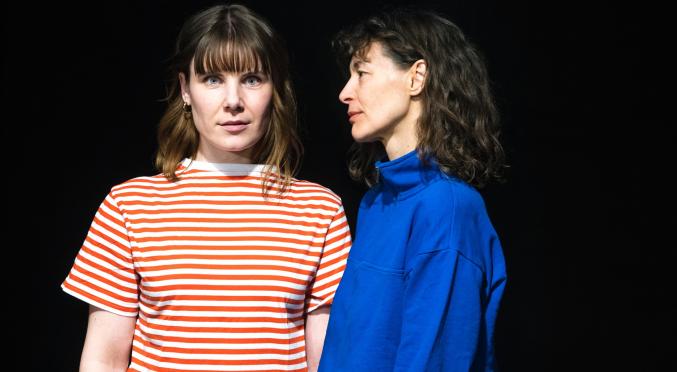 Zwei Frauen vor schwarzem Hintergrund: Eine mit rot-weiß gestreiftem T-Shirt und Pony, eine mit dunkelblauem Rollkragenpullover und lockigem Haar, beide leicht seitlich zueinander stehend.