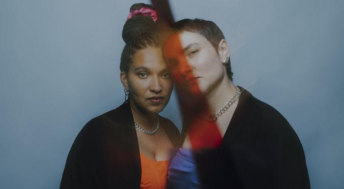 Zwei Personen eng beieinander: Eine mit hochgesteckten Haaren, pinkem Haarreif und silberner Kette, eine mit kurzem Haar und Kette, vor blauem Hintergrund mit farbigem Lichteffekt.